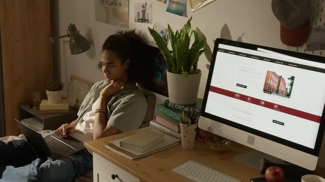 Handheld shot of young Black male student sitting on bed, typing on laptop and thinking while studying in bedroom. For online education, e-learning platforms, and student lifestyle promos