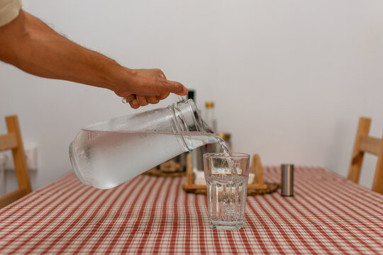 Hand pouring cold water from glass pitcher into drinking glass at home