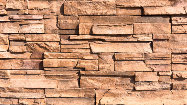 a full frame close view displays a textured tan stone wall constructed from flat rectangular pieces arranged in irregular horizontal rows creating layered shadows on the surface