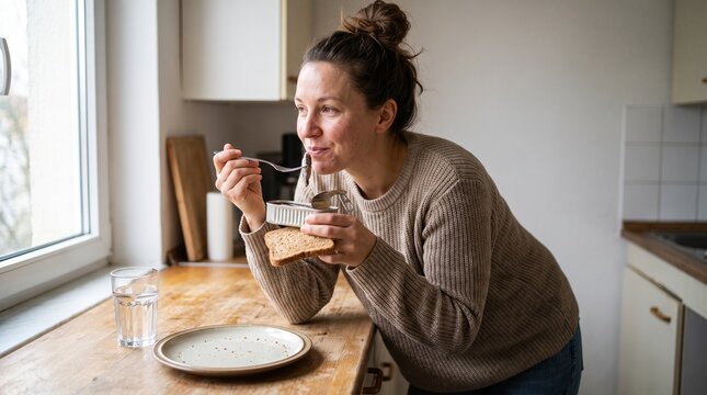 Cheerful young woman calmly eating canned fish and bread against a cozy kitchen background