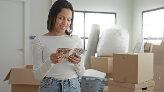 Woman counting mexican pesos with hands visible and smiling in a building packed with moving boxes and household items; moving day optimism.