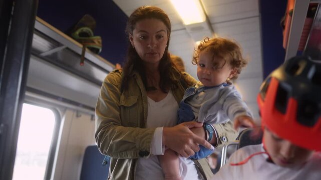 Mother carrying toddler beside boy with kick scooter inside passenger train before disembarking, capturing family travel, sibling bond and everyday public transportation