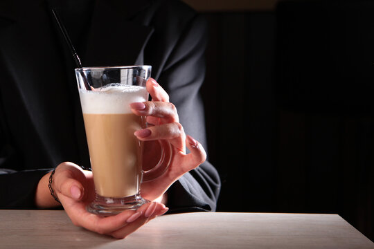 Beautiful hands are holding tall glass with coffee drink. Girl drinks latte. Latte coffee with foaming milk in tall glass. Copy space. Dark background