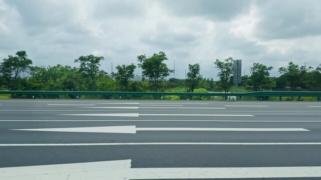 Empty asphalt highway with green metal guardrail and lush trees under a cloudy sky.