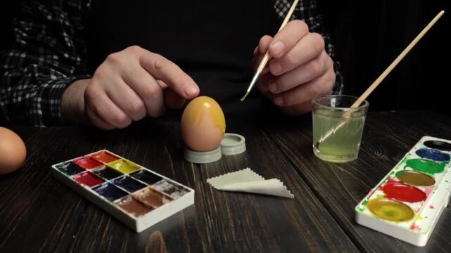 Person painting a decorative egg with a brush, surrounded by watercolor paints, eggs, and a glass of water on a wooden table
