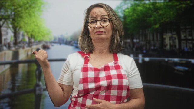 Woman with grey hair and glasses wearing red checkered apron points thumb backward at canal bridge in city; wry skepticism.