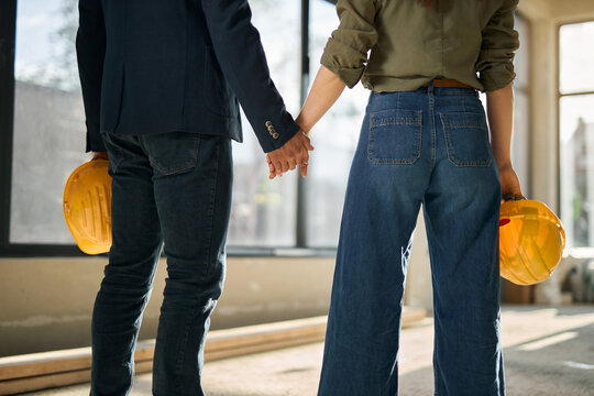 Rear view of a young couple holding hands and carrying a hardhat, looking at their apartment under construction