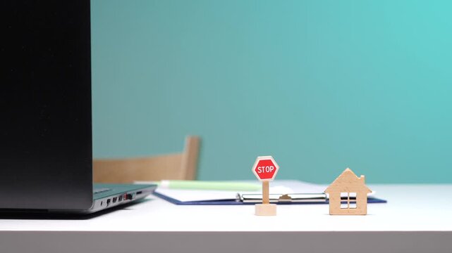 Hand placing a miniature stop sign next to a small wooden house model on a desk, symbolizing a halt to real estate transactions, eviction, or housing market difficulties