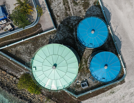Aerial view of three large circular storage tanks, one green and two blue, situated on a sandy ground area with a stone wall boundary in Vashafaru, Haa Alif Atoll, Maldives.