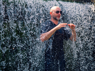Fototapeta premium Man stands under falling water. Water sprays around him. He holds hand out. Water droplets catch sunlight. He wears dark clothes. Background shows greenery. Scene shows natural water feature
