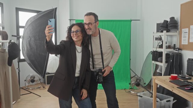 Woman and man photographers smile as woman holds smartphone and man holds camera strap while they review images in studio; creative teamwork joy.