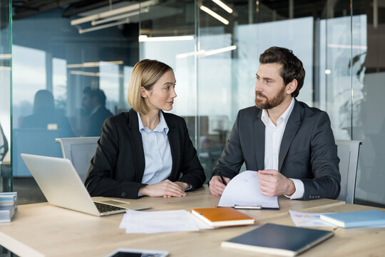 Two business professionals, a woman and a man, interacting and discussing work at a desk in a modern corporate office environment
