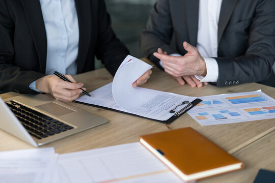 Business professionals reviewing agreement terms and discussing important financial details while signing a significant corporate contract on a wooden desk with a laptop and documents