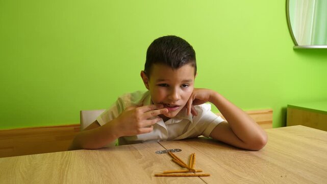 A boy sits at a table eating a breadstick snack while quietly enjoying a light meal in a bright room with green walls.