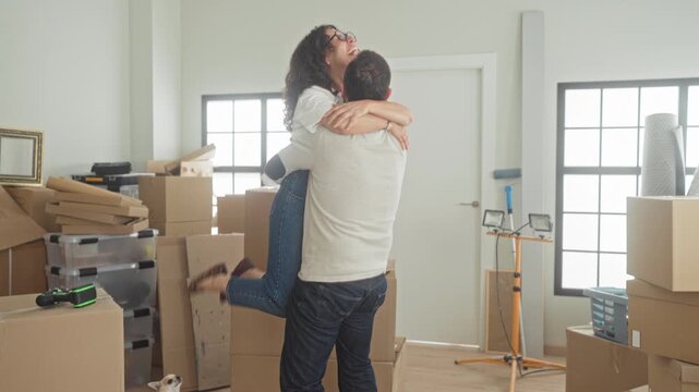 Man lifts woman in a tight hug amid stacked moving boxes, plastic bins, a work light and framed print in a building; moving day home joy.