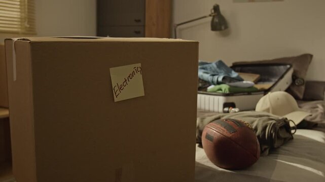 No people closeup shot of cardboard box labeled with sticky note sitting in messy bedroom next to open suitcase and football. Clip is perfect for student relocation, moving services, dorm life