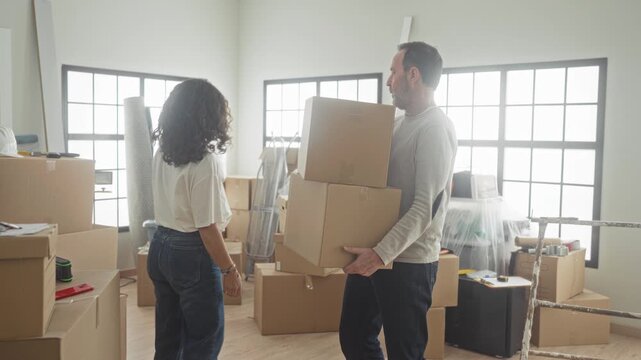 Man carrying stacked cardboard boxes while woman stacks boxes inside building during move; relocation teamwork hopeful.