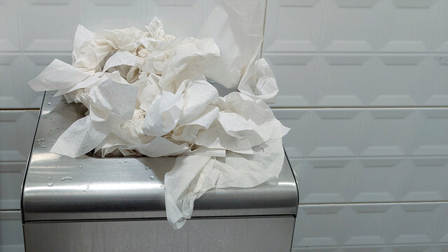 Overflowing waste bin full of crumpled paper hand towels in a public restroom blocking access, signifying a lack of hygiene, poor maintenance, and environmental neglect