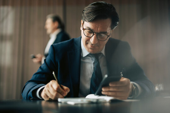 Mature businessman holding smart phone while writing in diary at office