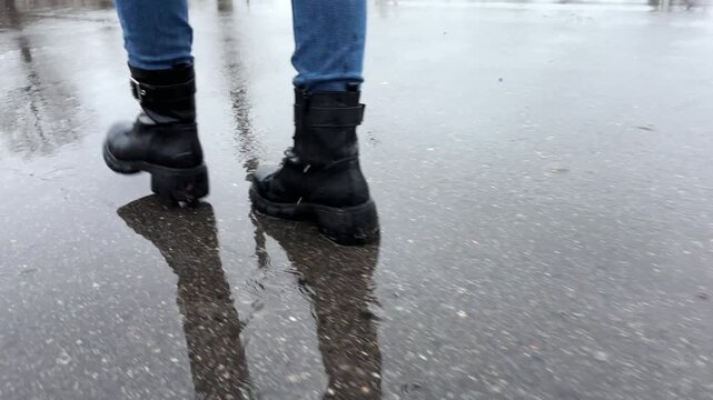 Female feet in boots going on wet asphalt road at spring or autumn. Legs of young woman stepping on puddles at rainy day. Concept of tranquility scene. Slow motion