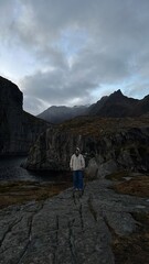 Fototapeta premium Female traveler standing on rocky cliffs in the Lofoten Islands, Norway. Dramatic mountain landscape under cloudy skies, ideal for travel and adventure concepts.