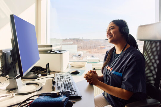 Smiling female doctor with hands clasped doing telehealth consultation on video call in clinic office