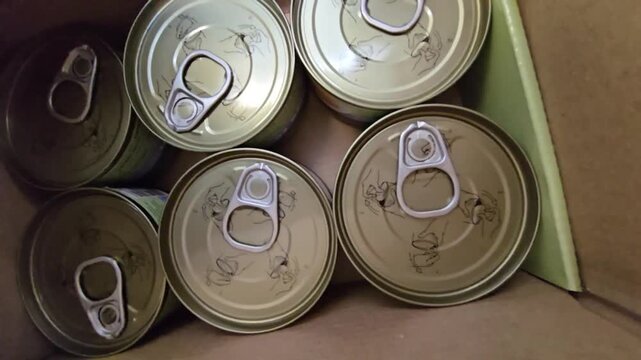 Top-down view of several sealed food cans with pull-tab lids inside a cardboard box.