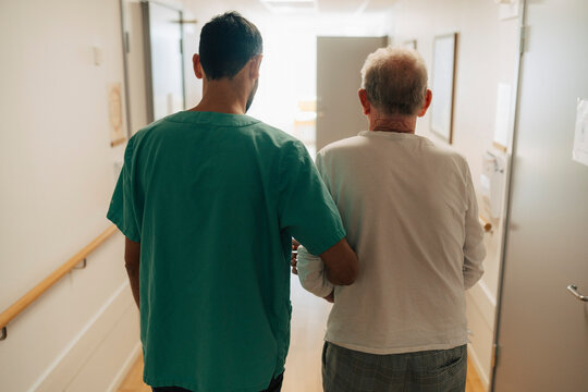 Rear view of male caregiver helping senior man walking in corridor