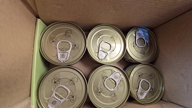 Top-down view of several sealed food cans with pull-tab lids inside a cardboard box.