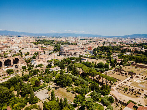 Aerial view of the Roman Forum with the Colosseum in the background, surrounded by lush green trees and historic ruins under a clear blue sky in Rome, Lazio, Italy.