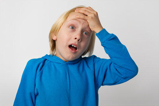 Young blond boy with wide blue eyes and an open mouth making a facepalm gesture, representing concepts of mistake, surprise, forgetfulness, or suddenly remembering something important