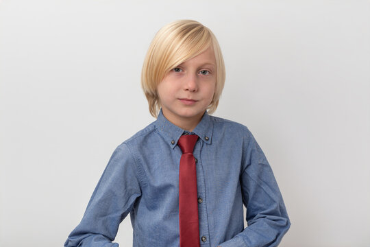 Blond boy standing against a white background, wearing a blue denim shirt and a red tie, representing concept of youth professionalism and smart attire
