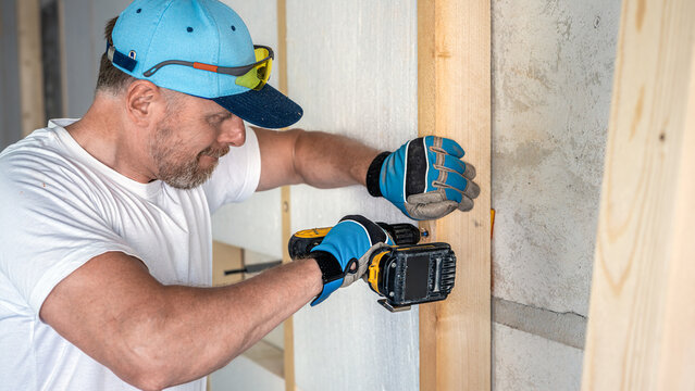 A worker uses an electric screwdriver for fixing a wooden prism to the wall using special screws with dowels. Dry construction of an insulated wall.
