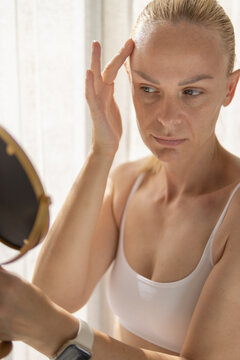 Woman scrutinizing her forehead for wrinkles in a mirror, focusing on skin problems and the natural aging process, considering dermatology and anti-aging skincare products