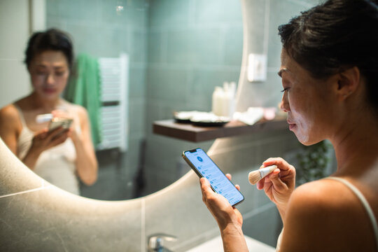 Woman applying makeup while using smartphone in home bathroom