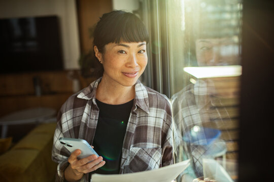 Smiling woman with smartphone and paperwork at home by window
