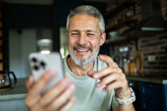Smiling middle-aged man video chatting on smartphone at home kitchen