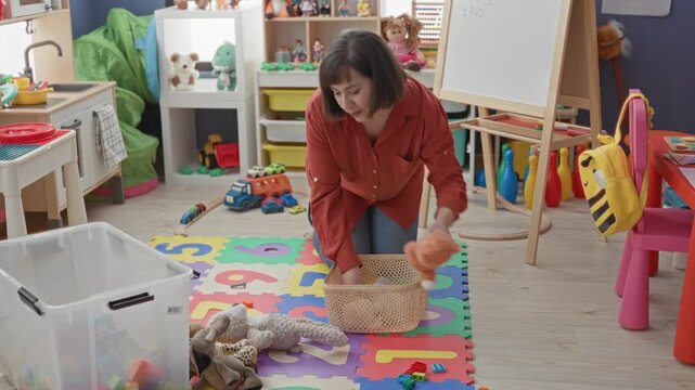 Woman kneeling and placing stuffed toys into a basket on a colorful number playmat in a preschool classroom; nurturing calm caregiving.