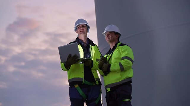 Engineers wearing safety helmets and jackets inspecting a wind turbine at wind farm at sunset. Concept renewable energy, sustainable technology wind power maintenance and environmental engineering.