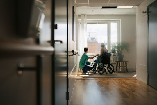 Female nurse spending leisure time with senior woman sitting in wheelchair near window