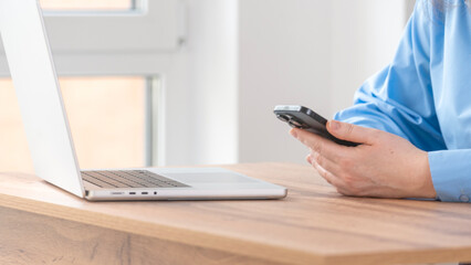 Fototapeta premium Close-up of person's hands holding a smartphone. Silver laptop rests on wooden desk surface. Light streams through window in background. Neutral setting captures modern work environment