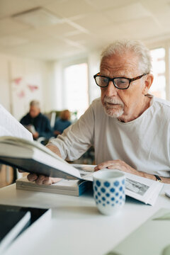 Focused senior man multi-tasking while reading books sitting in elder care facility