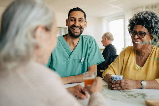 Happy male nurse interacting with senior women while sitting around table in retirement home