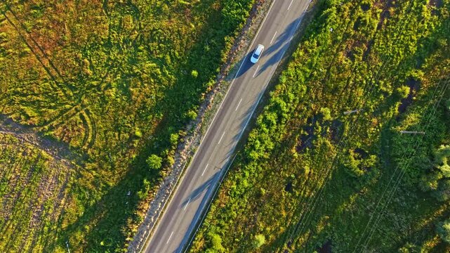 A road cuts through fields and forest areas under bright sunlight. Shadows form patterns on the green landscape. The scene shows nature and human-made structures coexisting.