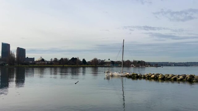 A sailboat sits moored beside a rock breakwater at English Bay, the domed Space Centre and downtown towers behind calm winter water under an overcast sky
