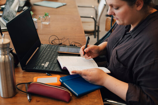 Female freelancer taking down notes in diary while sitting with laptop at shared working desk