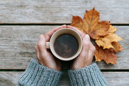 Hands cradle warm coffee cup. Crisp autumn leaves rest beside it. Cozy sweater sleeves frame the scene. Wooden surface adds rustic charm. Soothing moment feels both quiet and warm