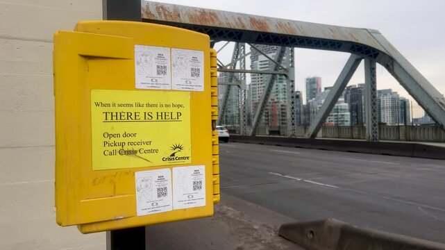 A yellow Crisis Centre phone box reading There is Help is mounted on the Burrard Bridge, steel truss and downtown towers beyond as traffic passes on a grey day