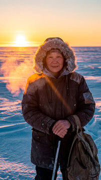 Weathered senior woman in fur hood parka standing on frozen sea at arctic sunrise
