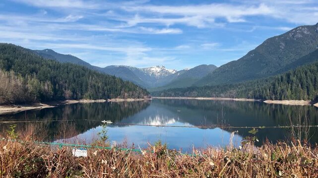 Snow-capped peaks reflect in the calm Capilano Reservoir above Cleveland Dam in North Vancouver, a log boom crossing the mirror-still water under a bright sky.
